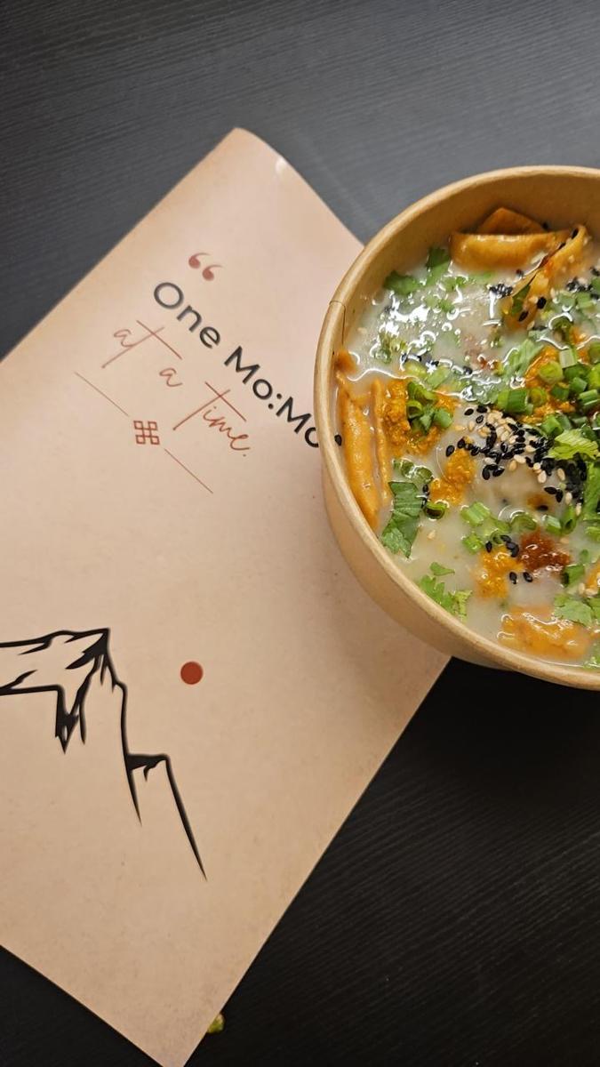 Mustang Jhol momos in broth with sesame, spring onion and chilli, beside the kraft menu reading One Mo:Mo at a time