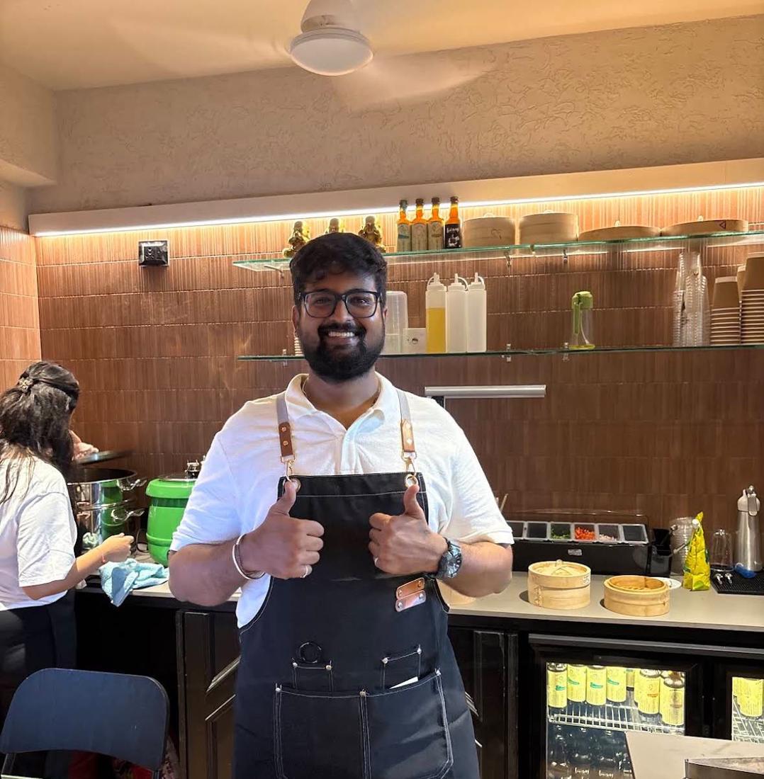 Aman Kapadia in a Mustang apron, giving two thumbs up behind the café counter